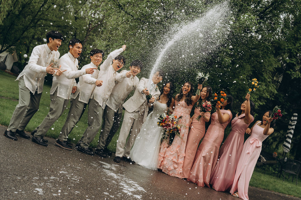 A bride and groom pop a bottle of champagne while their bridal party celebrates next to them.
